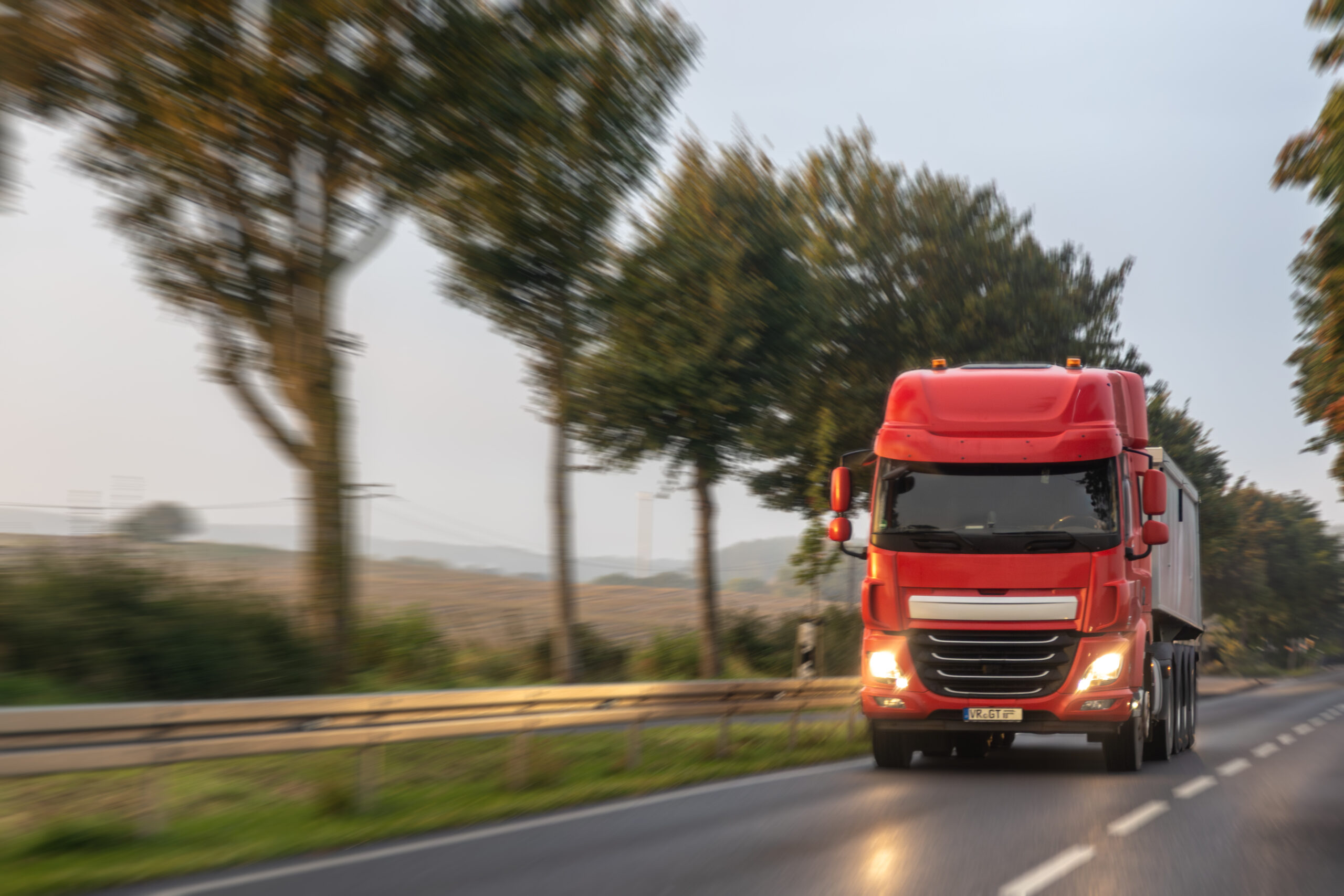 A red tractor unit moves along a country highway between trees, gentle morning light shaping the cabin. The feel of schedule, route and on time service.